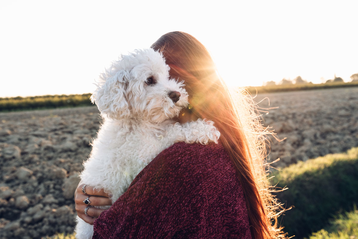 a fotón egy hosszú hajú fiatal nő a vállára vette fehér szőrű kutyusát, és a szabadban egy szántóföld mellett állnak